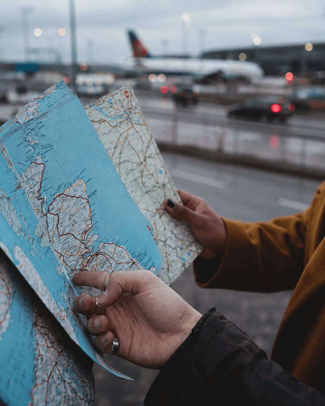 Two hands holding an open folded road map, with an airport apron and a parked aircraft soft-focused behind.